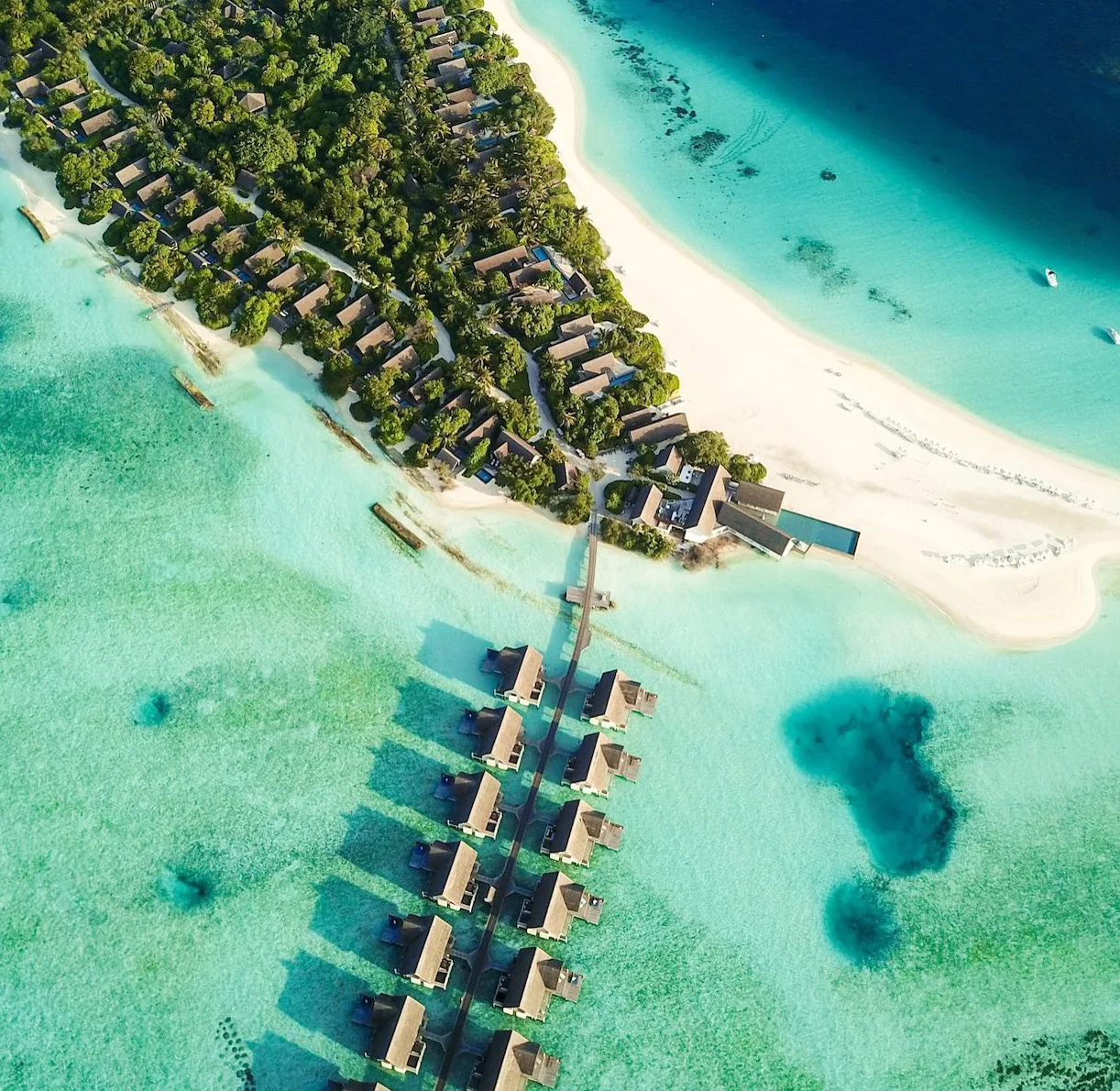 Aerial photograph of Four Seasons Landaa Giraavaru: a curving jetty of over-water villas over turquoise lagoon beside a wooded island.